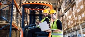 Two warehouse workers at Radical Cosmetics stand side by side in a clean, well-lit facility, comparing notes on a clipboard. One wears a navy-blue uniform while the other is dressed in a charcoal-gray shirt, both donning protective gloves and hairnets to ensure GMP compliance. Surrounded by stacked boxes of private label cosmetics and skincare products, the workers review inventory and batching details as part of quality control. Keywords: cosmetics warehouse operations, private label beauty manufacturing, GMP-compliant facility, skincare production, color cosmetics logistics, eco-friendly cosmetic packaging, clean beauty supplier, inventory management, contract cosmetic manufacturing.