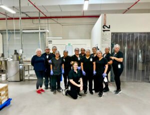 Radical Cosmetics production team standing in front of Production Line 12, where solid and powder cosmetic manufacturing takes place. The image features lab technicians, batching specialists, and production line workers in clean lab coats and protective gear, set against a backdrop of high-grade stainless steel equipment. Showcasing our commitment to clean beauty, high-quality cosmetic production, and USA-made manufacturing of paraben-free, cruelty-free, and non-toxic makeup and skincare products.